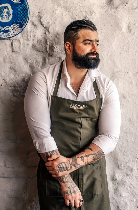 Paco Ruano, chef de Alcalde, con camisa blanca y mandil, frente a una pared de azulejos y yeso.
